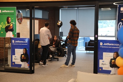 A modern office setting features several people engaging in conversation. Posters advertising Jotform SIGN are prominently displayed on the walls and partitions. The space includes brick walls and large windows, with some natural light coming through. Balloons are in the background, adding a celebratory or casual atmosphere. The individuals are casually dressed, lending a relaxed tone to the environment.