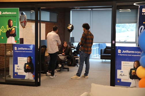 A modern office setting features several people engaging in conversation. Posters advertising Jotform SIGN are prominently displayed on the walls and partitions. The space includes brick walls and large windows, with some natural light coming through. Balloons are in the background, adding a celebratory or casual atmosphere. The individuals are casually dressed, lending a relaxed tone to the environment.