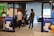 A modern office setting features several people engaging in conversation. Posters advertising Jotform SIGN are prominently displayed on the walls and partitions. The space includes brick walls and large windows, with some natural light coming through. Balloons are in the background, adding a celebratory or casual atmosphere. The individuals are casually dressed, lending a relaxed tone to the environment.