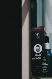 A bottle of beard growth oil with a dropper cap is positioned in front of its packaging box. The bottle and box are adorned with branding and text indicating the product's purpose. The scene has a moody ambiance due to the lighting and shadows.