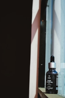 A bottle of beard growth oil rests on a window ledge. The packaging is dark with a metallic cap, and it features clear labeling. The background shows contrasting light and shadow formed by sunlight streaming through the window.