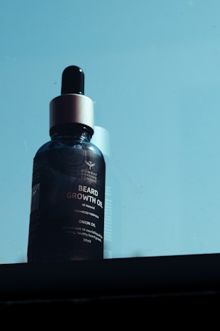 A bottle of beard growth oil with a dropper cap is set against a light blue background. The label on the bottle prominently displays the brand and product information.