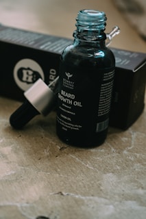 A bottle of beard growth oil with a dropper is placed on a textured surface. The label is visible, indicating the brand and product details. The packaging box is partially seen in the background.