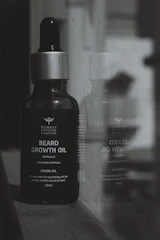 A bottle of Bombay Shaving Company beard growth oil is positioned next to a reflective surface, showing its reflection. The bottle is dark with a silver cap and is labeled with details about the product, including 'Onion Oil' and '30ml'.