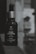 A bottle of Bombay Shaving Company beard growth oil is positioned next to a reflective surface, showing its reflection. The bottle is dark with a silver cap and is labeled with details about the product, including 'Onion Oil' and '30ml'.