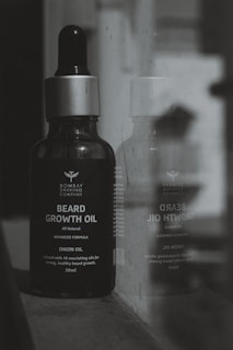 A bottle of Bombay Shaving Company beard growth oil is positioned next to a reflective surface, showing its reflection. The bottle is dark with a silver cap and is labeled with details about the product, including 'Onion Oil' and '30ml'.