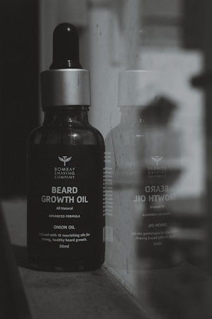 A bottle of Bombay Shaving Company beard growth oil is positioned next to a reflective surface, showing its reflection. The bottle is dark with a silver cap and is labeled with details about the product, including 'Onion Oil' and '30ml'.