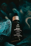A bottle of beard growth oil is displayed in the center, surrounded by a textured blue netting. The oil bottle features a label with text that describes the contents as 'Beard Growth Oil' with onion oil and nourishing attributes. The lighting is moody and dark, with shadows cast by the netting.