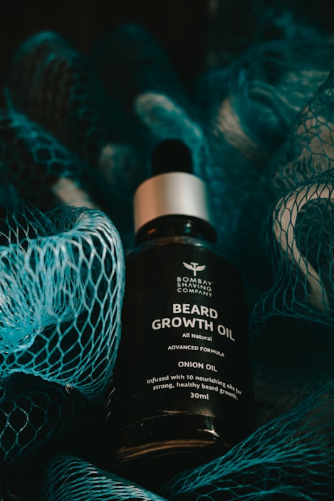 A bottle of beard growth oil is displayed in the center, surrounded by a textured blue netting. The oil bottle features a label with text that describes the contents as 'Beard Growth Oil' with onion oil and nourishing attributes. The lighting is moody and dark, with shadows cast by the netting.