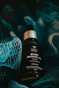 A bottle of beard growth oil is displayed in the center, surrounded by a textured blue netting. The oil bottle features a label with text that describes the contents as 'Beard Growth Oil' with onion oil and nourishing attributes. The lighting is moody and dark, with shadows cast by the netting.