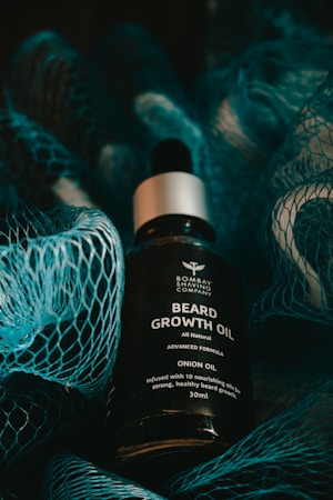 A bottle of beard growth oil is displayed in the center, surrounded by a textured blue netting. The oil bottle features a label with text that describes the contents as 'Beard Growth Oil' with onion oil and nourishing attributes. The lighting is moody and dark, with shadows cast by the netting.