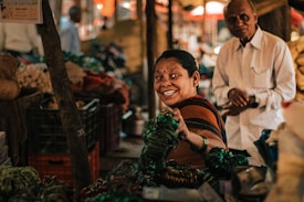 A woman with a warm smile holds green bangles in a bustling market setting. A man stands nearby, partially in the frame. The market is filled with various goods and people are visible in the background, creating a lively atmosphere.