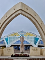 A modern architectural structure features a large, arching gateway in the foreground with star motifs. In the background, a dome-like building displays vibrant colors, including yellow, blue, and white geometric patterns. There is a central sculptural element in front of the entrance, and the words on the signs suggest it is a cultural or historical center.