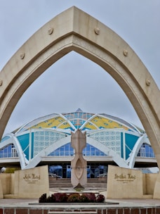 A modern architectural structure features a large, arching gateway in the foreground with star motifs. In the background, a dome-like building displays vibrant colors, including yellow, blue, and white geometric patterns. There is a central sculptural element in front of the entrance, and the words on the signs suggest it is a cultural or historical center.