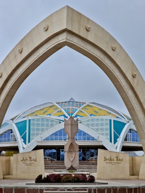 A modern architectural structure features a large, arching gateway in the foreground with star motifs. In the background, a dome-like building displays vibrant colors, including yellow, blue, and white geometric patterns. There is a central sculptural element in front of the entrance, and the words on the signs suggest it is a cultural or historical center.