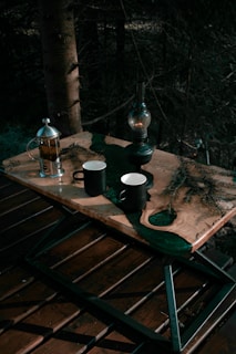 A wooden table outdoors with two black mugs, one of which is partially filled, a metallic French press containing tea or coffee, and an antique oil lamp. Pine branches are scattered across the table, adding to the rustic, natural setting amid a dark forest background.