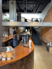 A modern cafe interior with a curved wooden counter and industrial design elements. The counter showcases baked goods under a glass display, and there is coffee-making equipment including an espresso machine. A blurred figure is seen behind the counter, suggesting movement. The space includes exposed brick walls, concrete surfaces, and a staircase leading to an upper level.