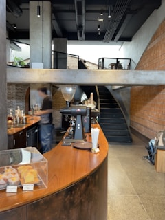 A modern cafe interior with a curved wooden counter and industrial design elements. The counter showcases baked goods under a glass display, and there is coffee-making equipment including an espresso machine. A blurred figure is seen behind the counter, suggesting movement. The space includes exposed brick walls, concrete surfaces, and a staircase leading to an upper level.
