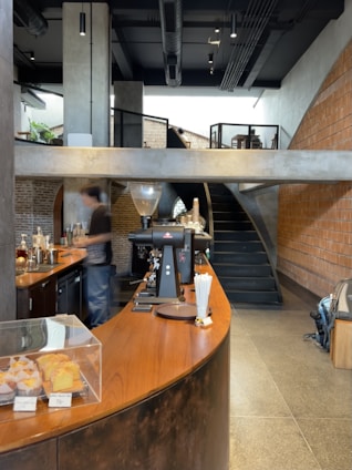A modern cafe interior with a curved wooden counter and industrial design elements. The counter showcases baked goods under a glass display, and there is coffee-making equipment including an espresso machine. A blurred figure is seen behind the counter, suggesting movement. The space includes exposed brick walls, concrete surfaces, and a staircase leading to an upper level.