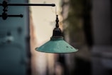 An old, vintage green metal lamp hangs from a horizontal pipe against a blurred city street background. The lamp shows signs of rust and wear, adding to its aged appearance.