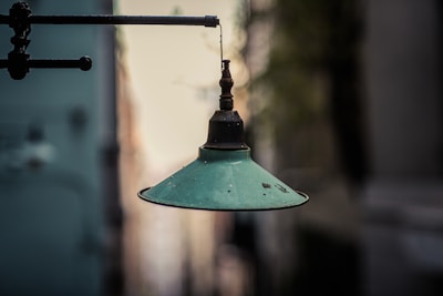 An old, vintage green metal lamp hangs from a horizontal pipe against a blurred city street background. The lamp shows signs of rust and wear, adding to its aged appearance.