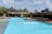A swimming pool is surrounded by greenery and wooden structures, creating a serene outdoor space. Flags from various countries are strung across in vibrant colors, adding an international flair. There are colorful cushions placed poolside, providing a casual lounging area. A sign with the logo 'Jotform' is prominently displayed, suggesting a branded event or setting.