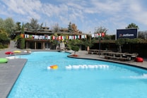 A swimming pool is surrounded by greenery and wooden structures, creating a serene outdoor space. Flags from various countries are strung across in vibrant colors, adding an international flair. There are colorful cushions placed poolside, providing a casual lounging area. A sign with the logo 'Jotform' is prominently displayed, suggesting a branded event or setting.
