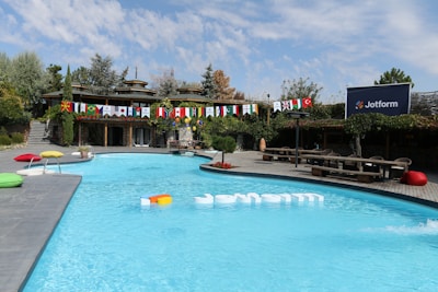 A swimming pool is surrounded by greenery and wooden structures, creating a serene outdoor space. Flags from various countries are strung across in vibrant colors, adding an international flair. There are colorful cushions placed poolside, providing a casual lounging area. A sign with the logo 'Jotform' is prominently displayed, suggesting a branded event or setting.
