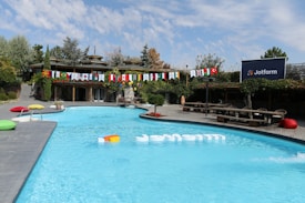 A swimming pool is surrounded by greenery and wooden structures, creating a serene outdoor space. Flags from various countries are strung across in vibrant colors, adding an international flair. There are colorful cushions placed poolside, providing a casual lounging area. A sign with the logo 'Jotform' is prominently displayed, suggesting a branded event or setting.