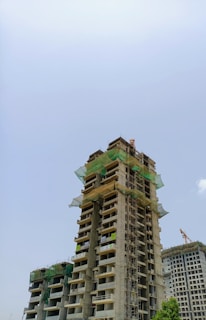 A tall concrete building under construction, featuring green netting and scaffolding around various sections. The structure has multiple floors with open spaces for windows and balconies, and a crane is visible in the background. The sky is clear with a light blue hue.