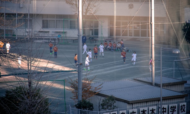 Children are playing a soccer game on a fenced outdoor field. The players are wearing different colored uniforms, indicating opposing teams. Some children are seated on the sidelines watching the game. The surroundings include a building with large windows and some trees.