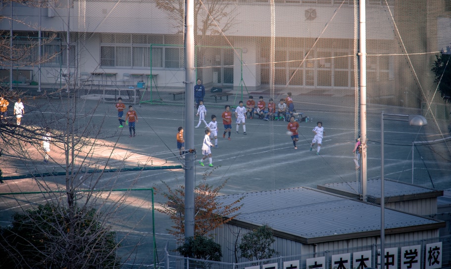 Children are playing a soccer game on a fenced outdoor field. The players are wearing different colored uniforms, indicating opposing teams. Some children are seated on the sidelines watching the game. The surroundings include a building with large windows and some trees.