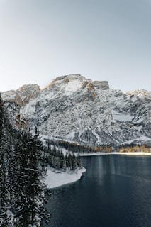 A serene mountain landscape with snow-capped peaks rising above a calm lake. Evergreen trees line the shore, and the reflection of the mountains can be faintly seen in the water. The sky is clear with a subtle gradient from light to darker tones.