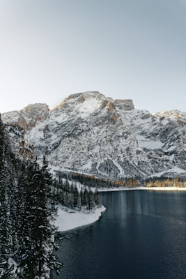 A serene mountain landscape with snow-capped peaks rising above a calm lake. Evergreen trees line the shore, and the reflection of the mountains can be faintly seen in the water. The sky is clear with a subtle gradient from light to darker tones.