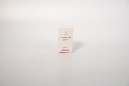 A small rectangular box with a white background, displaying text related to acne treatment. The box has pink text on the front that includes the words 'Treatment Acne'. The surface is smooth and the background is a neutral, light cream color.