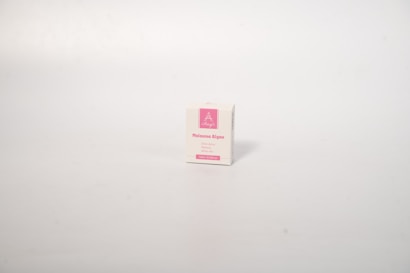 A small white box with pink accents stands isolated on a plain light background. The box appears to be a product package with text indicating 'Melasma Algae' and other details in pink text.