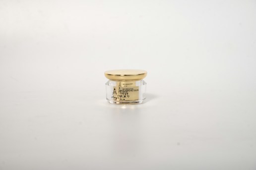 A small, transparent cosmetic jar with a golden lid, positioned in the center against a plain white background. The jar contains a cream and has black text on it detailing the product's name and features.