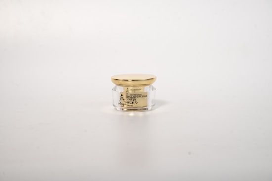 A small, transparent cosmetic jar with a golden lid, positioned in the center against a plain white background. The jar contains a cream and has black text on it detailing the product's name and features.