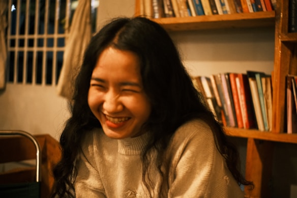 A person with long dark hair smiles broadly while sitting in a cozy, warmly lit room. Shelves filled with various books are visible in the background, adding a sense of comfort and relaxation to the setting.