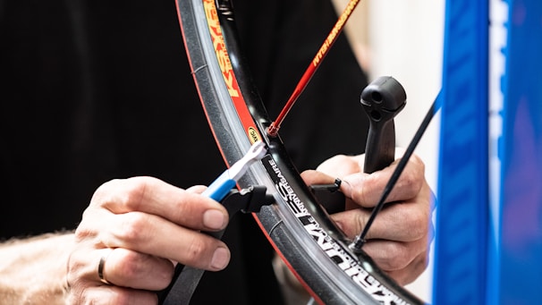 Hands are working on a bicycle wheel, focusing on adjusting the spokes with a tool. The tire branding is visible, and the setting appears to be a repair or workshop environment.