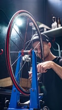 A person in a workshop is adjusting a bicycle wheel held in a blue wheel truing stand. The focus is on the intricate components of the wheel, including the spokes and tire labeled with branding. The individual appears to be concentrating on precision work, illuminated by a bright lamp overhead. Shelves and tools are visible in the dimly lit background, adding to the workshop atmosphere.