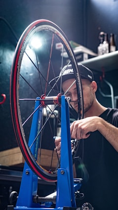 A person in a workshop is adjusting a bicycle wheel held in a blue wheel truing stand. The focus is on the intricate components of the wheel, including the spokes and tire labeled with branding. The individual appears to be concentrating on precision work, illuminated by a bright lamp overhead. Shelves and tools are visible in the dimly lit background, adding to the workshop atmosphere.