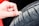 A close-up of a hand using a coin to check the tread depth of a car tire. The tire appears to be new or in good condition, and the background suggests the vehicle might be red.