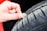 A close-up of a hand using a coin to check the tread depth of a car tire. The tire appears to be new or in good condition, and the background suggests the vehicle might be red.