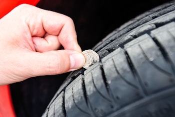 A close-up of a hand using a coin to check the tread depth of a car tire. The tire appears to be new or in good condition, and the background suggests the vehicle might be red.
