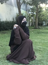 A person wearing a brown abaya sits on green grass in an outdoor setting. The background includes trees and a building near the person, suggesting a garden or park environment. The individual is looking to the side and wears a black hijab and face covering.