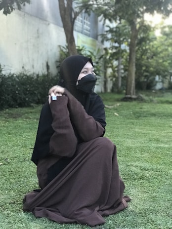 A person wearing a brown abaya sits on green grass in an outdoor setting. The background includes trees and a building near the person, suggesting a garden or park environment. The individual is looking to the side and wears a black hijab and face covering.