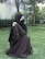 A person wearing a brown abaya sits on green grass in an outdoor setting. The background includes trees and a building near the person, suggesting a garden or park environment. The individual is looking to the side and wears a black hijab and face covering.