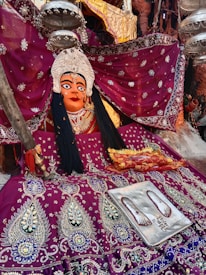 A vibrant and ornate statue draped in an intricately embroidered magenta garment adorned with detailed patterns and embellishments. The statue has a striking face, accentuated with bold makeup and framed by ornate headgear. Silver ceremonial items hang nearby, complementing the rich textiles. A silver plate with religious symbols and painted footprints rests on the fabric, adding a sacred element to the scene.