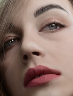 A close-up of a person's face focusing on well-defined eyebrows, expressive eyes with subtle makeup, and full lips adorned with a matte pink lipstick. The image captures fine details of the skin texture and hair strands.